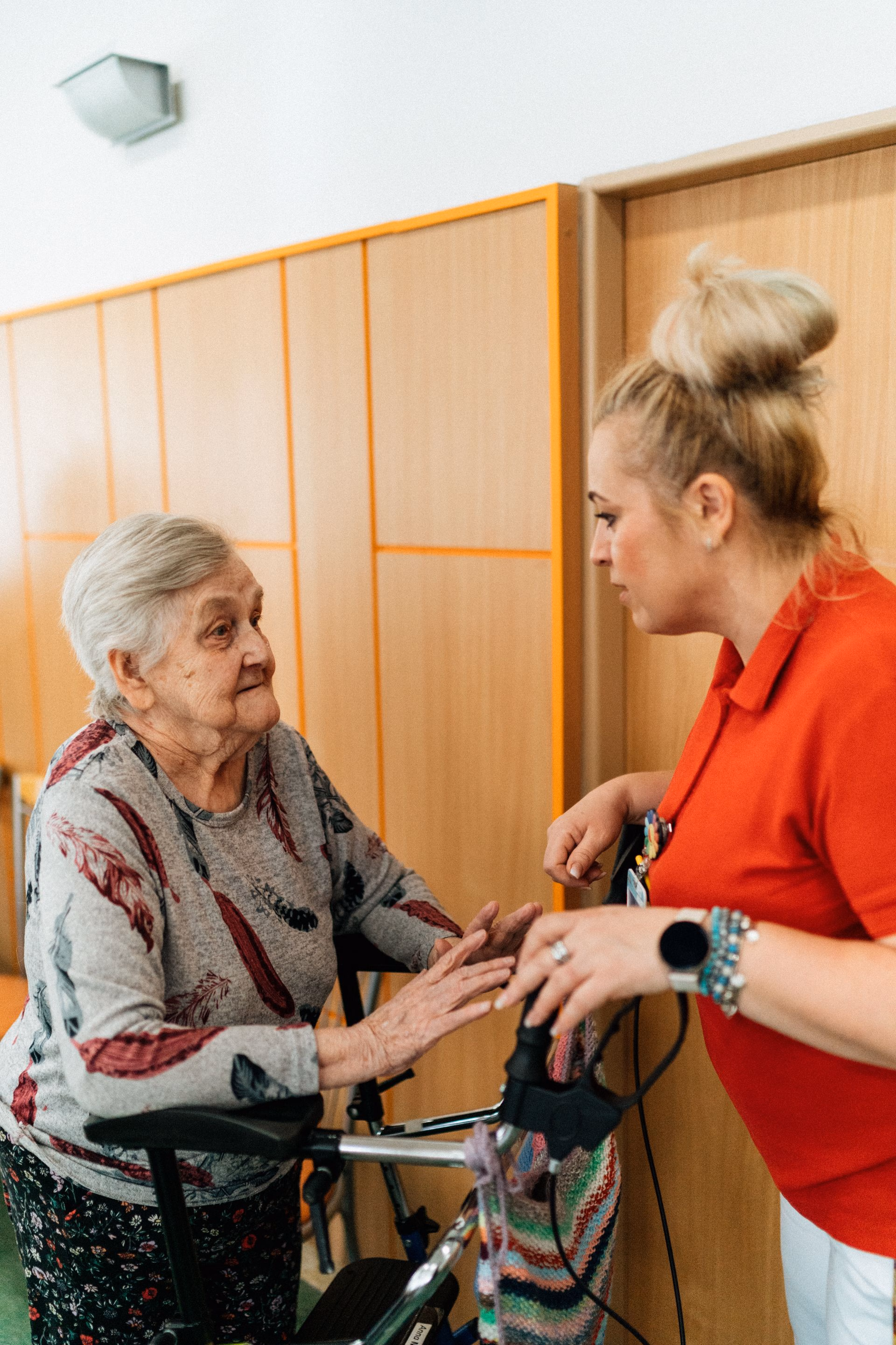 Elderly woman with a walker talking to a caregiver in a red shirt in a hallway.