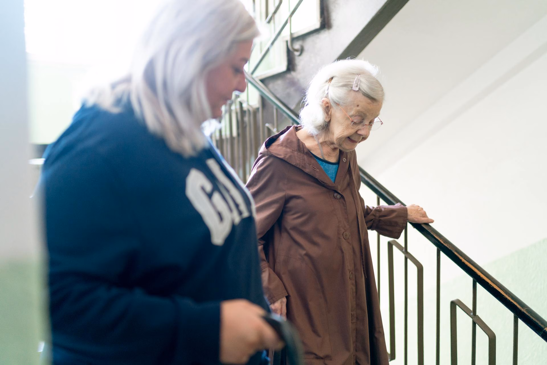 Elderly woman holding stair railing walking down stairs with younger woman in blue sweatshirt nearby.