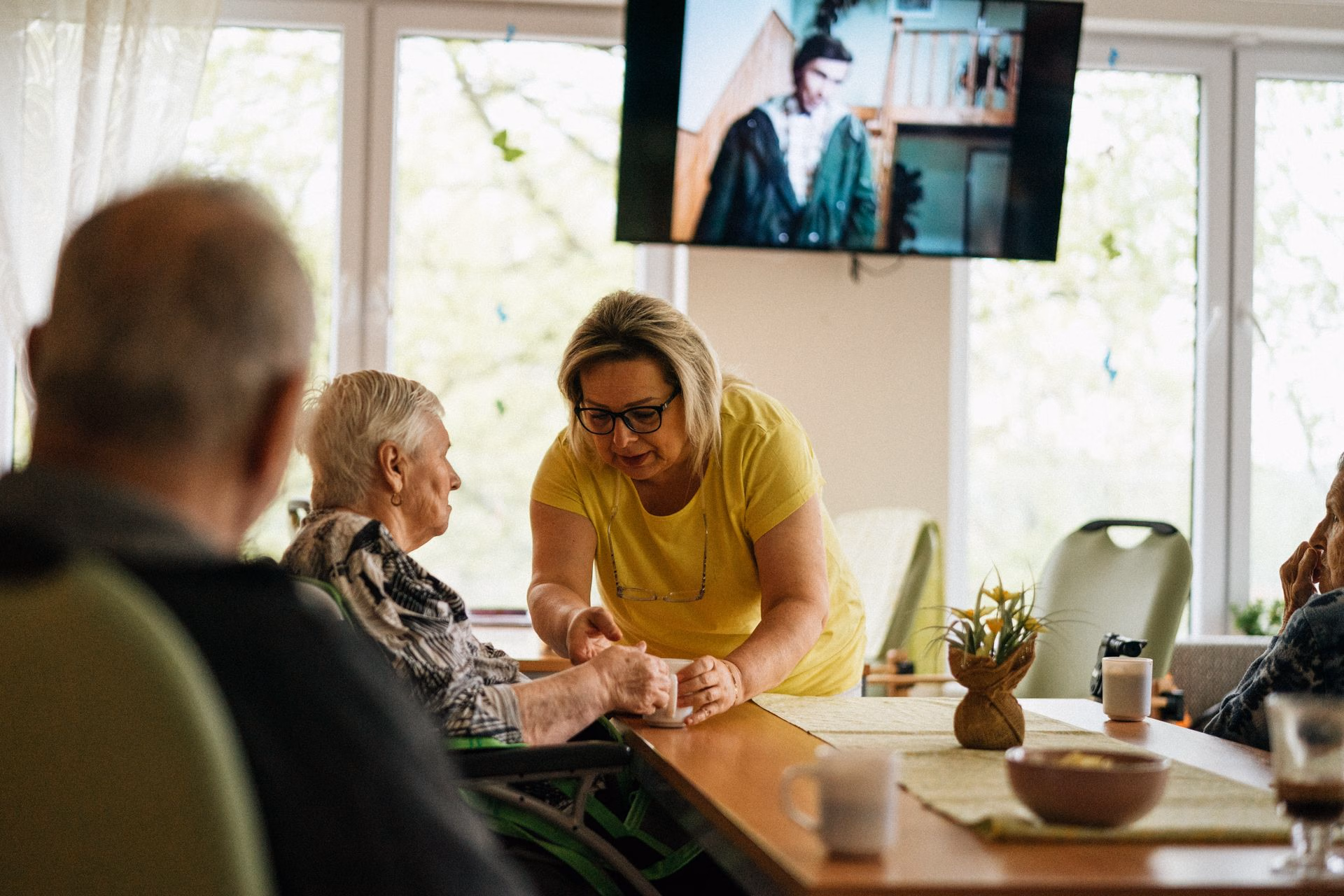 Woman in yellow shirt helping elderly woman in wheelchair by handing her a cup at a table.